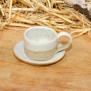 Op de afbeelding: Een kleine, handgemaakte keramische koffiekop en schotel set. De kop en schotel zijn gebroken wit met een beige band aan de basis. De kop heeft een afgerond oor. De set staat op een houten oppervlak met stro op de achtergrond.