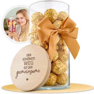Könnte beinhalten: Ein Glasgefäß gefüllt mit goldverpackten Pralinen, gebunden mit einem goldenen Band und Schleife. Das Gefäß hat einen Holzdeckel mit dem Text "Der schönste Weg ist der gemeinsame" eingraviert.