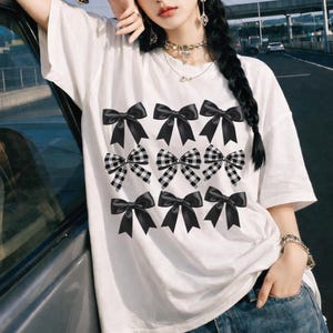 Puede incluir: Camiseta blanca con un diseño de lazos negros y lazos a cuadros blancos y negros. Los lazos están dispuestos en filas en la parte delantera. La camiseta es de estilo casual y holgado.