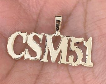 May include: Gold pendant necklace with the text "CSM51" in a bold, blocky font.