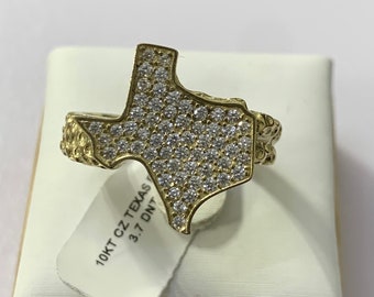 May include: A gold-colored ring in the shape of the state of Texas, encrusted with small, clear stones. The ring is on a white surface, with a tag that reads "10KT CZ Texas 3.7 DNT". The ring has a textured design on the band.