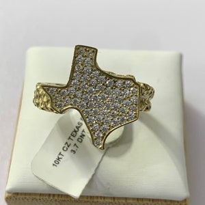 May include: A gold-colored ring in the shape of the state of Texas, encrusted with small, clear stones. The ring is on a white surface, with a tag that reads "10KT CZ Texas 3.7 DNT". The ring has a textured design on the band.