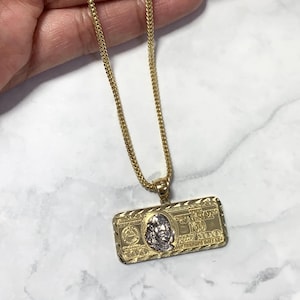 May include: A gold-toned necklace with a rectangular pendant shaped like a $100 bill. The pendant features a detailed design with a portrait and the number 100 in silver and gold tones. The chain is a fine, textured gold.