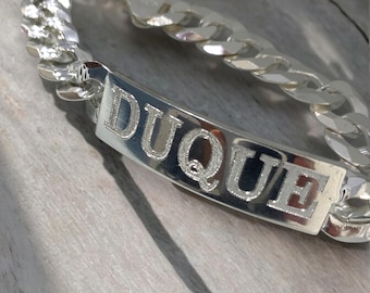 May include: Silver curb chain bracelet with a silver nameplate engraved with the name "DUQUE".