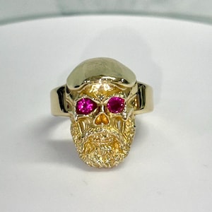 May include: A gold ring featuring a sculpted skull with a detailed beard and mustache. The skull has two vibrant pink circular gemstones set into the eye sockets, and the metal surface has a polished finish on the cranium and textured details.