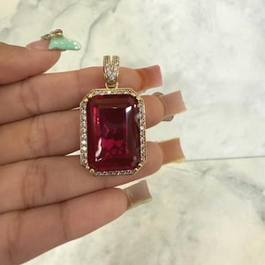 May include: A gold pendant with a large, rectangular, deep red gemstone. The gemstone is surrounded by a border of small, clear stones. The bail of the pendant is also adorned with clear stones. The pendant is held in a hand.