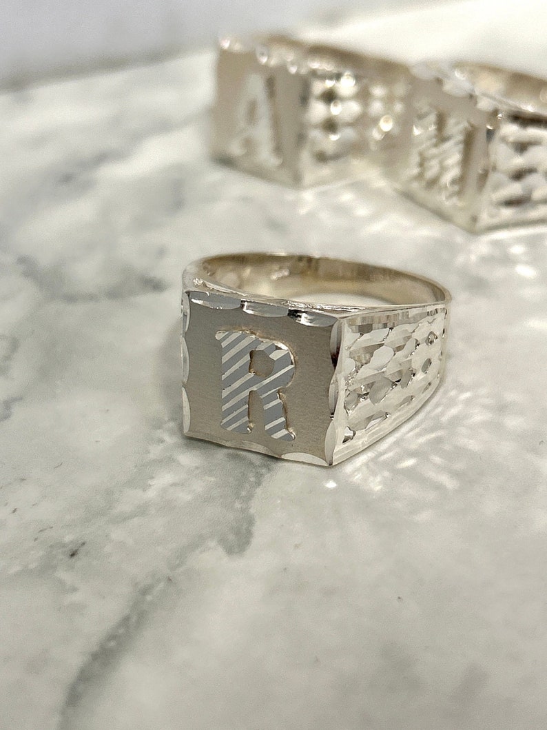 May include: Silver ring with a square face featuring the letter "R" in a raised, textured design. The ring has a textured band.