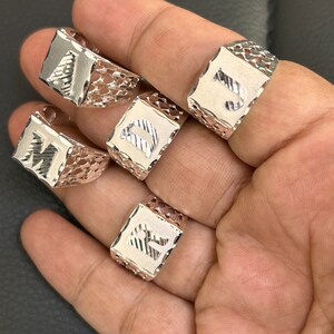 May include: Four silver rings with different letters engraved on them. The rings are square shaped and have a delicate, intricate design.