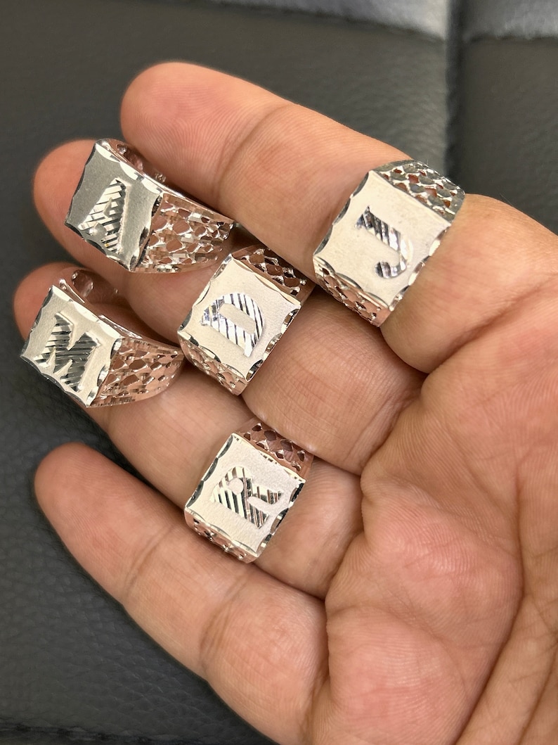 May include: Four silver rings with different letters engraved on them. The rings are square shaped and have a delicate, intricate design.