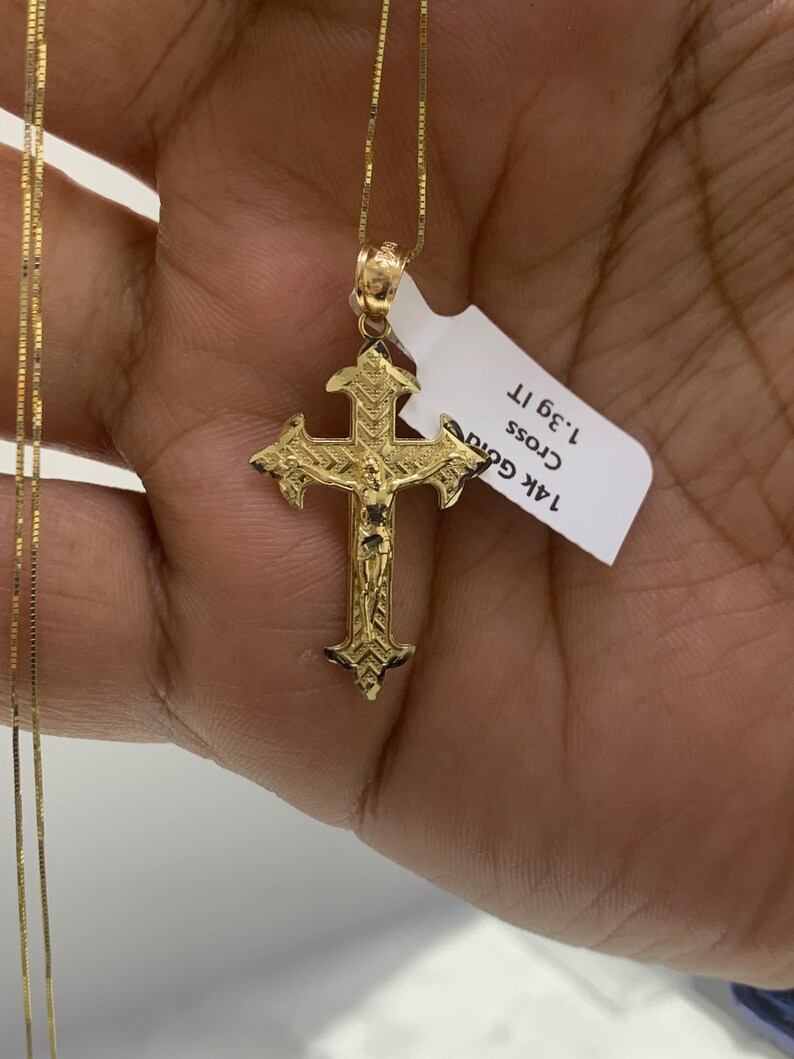 May include: A gold chain necklace with a detailed gold cross pendant. The pendant features a figure of Jesus Christ. The tag attached to the necklace reads "14k Gold Cross 1.3g 1T".
