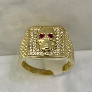 May include: A gold-colored ring featuring a skull design. The skull has red gemstone eyes and is set within a square frame encrusted with small, clear gemstones. The ring band has a textured pattern.
