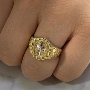 May include: A gold ring with an oval-shaped setting featuring an ankh symbol. The ring has a textured band.