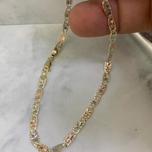 K&ouml;nnte beinhalten: Ein goldfarbenes Kettenarmband mit dreifarbigem Design. Die Kette besteht aus kleinen, ovalen Gliedern mit einem rautenf&ouml;rmigen Muster.