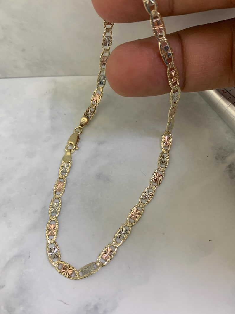 K&ouml;nnte beinhalten: Ein goldfarbenes Kettenarmband mit dreifarbigem Design. Die Kette besteht aus kleinen, ovalen Gliedern mit einem rautenf&ouml;rmigen Muster.