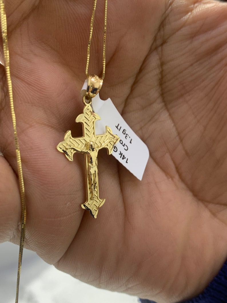 May include: A gold chain necklace with a detailed gold cross pendant. The pendant features a figure of Jesus Christ. The tag attached to the chain reads "14K Gold Cross 1.3g 1T."
