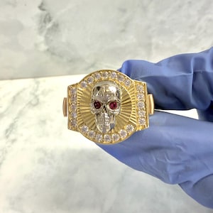 May include: A gold-toned ring featuring a skull design with red gemstone eyes. The skull is set in a silver-colored metal, surrounded by a sunburst pattern and encrusted with clear gemstones. The ring has a rectangular shape.