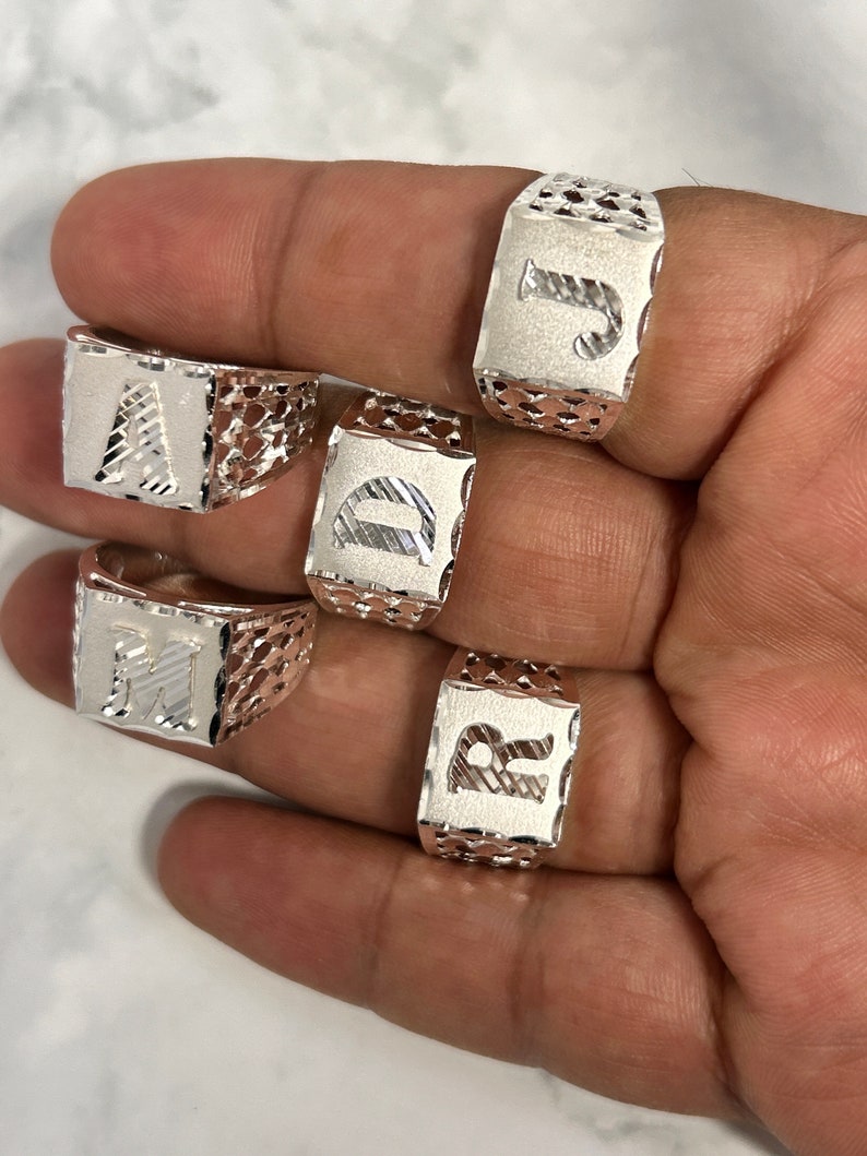 May include: Five silver rings with different letters engraved on them. The letters are A, J, D, M, and R. The rings are on a hand.