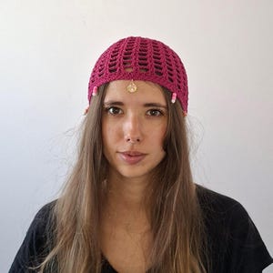 Puede incluir: Un gorro de ganchillo magenta con un colgante dorado y detalles de cuentas. El gorro tiene un diseño de tejido abierto y está adornado con pequeñas cuentas rosas y blancas. Un colgante dorado cuelga de la parte delantera del gorro.