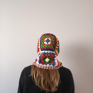 May include: A colourful crocheted hat with a granny square pattern in shades of red, orange, yellow, green, blue, and white. The hat is worn by a person with long brown hair.