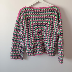 May include: A long-sleeved sweater crafted with a crochet granny square pattern in alternating rows of vibrant hot pink, deep forest green, and off-white yarn. The garment features a wide boat neckline and a straight hem, hanging on a white hanger.