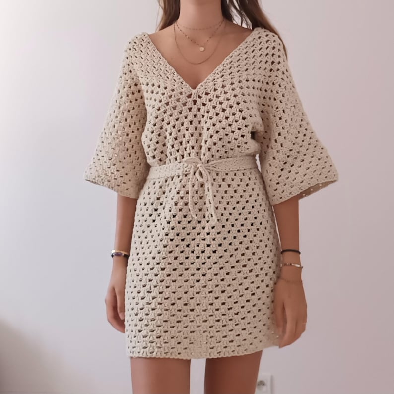 May include: A beige crochet dress with a V-neckline and a tie belt. The dress has a loose fit and is made of a lightweight, breathable fabric.