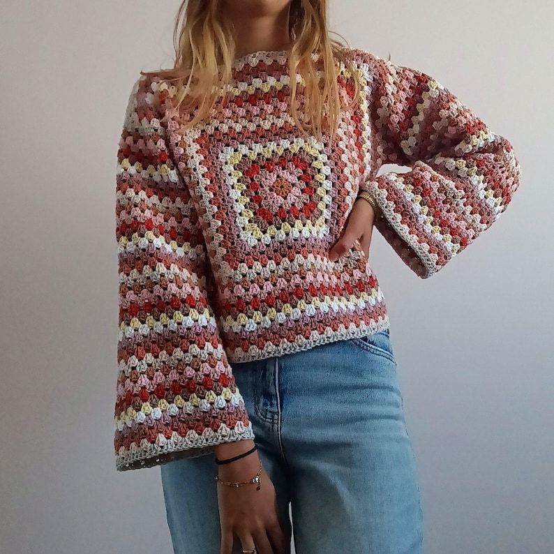 May include: A long-sleeved crochet sweater featuring a multi-colored granny square pattern in shades of red, pink, yellow, and white. The garment has a round neckline and wide, flared sleeves, paired with light blue denim jeans.