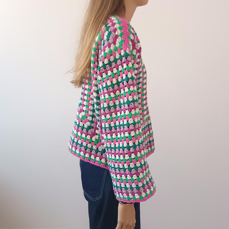 May include: A long-sleeved crochet cardigan featuring a vibrant granny square pattern in shades of hot pink, emerald green, white, and lavender. The garment has a boxy silhouette with a round neckline and is paired with dark blue denim jeans.