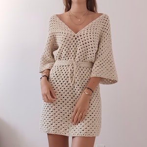 May include: A crocheted dress with a V-neck, 3/4 sleeves, and a tie belt. The dress is made of a light beige yarn and has a loose, flowy fit.