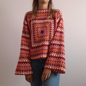 May include: A long-sleeved crochet sweater featuring a multi-colored granny square pattern in shades of red, orange, pink, and cream. The garment has a boxy fit with wide, flared sleeves and a round neckline, paired with blue denim jeans.