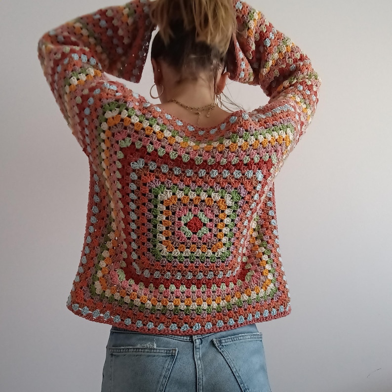 May include: A long-sleeved crochet sweater featuring a multi-colored granny square pattern in shades of coral, pink, green, yellow, and light blue. The garment has a wide neckline and a boxy fit, paired with light blue denim jeans.