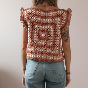 May include: A handmade crochet top in a granny square pattern. The top features a square neckline and ruffled sleeves. The color palette includes shades of brown, pink, and white. The top is paired with blue jeans.