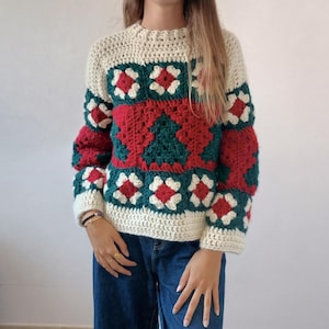 Könnte beinhalten: Ein cremefarbener Häkelpullover mit festlichem Design. Der Pullover hat rote, grüne und weiße quadratische und Baum-Motive. Der langärmlige Pullover ist aus einem strukturierten Garn gefertigt und hat einen runden Ausschnitt.
