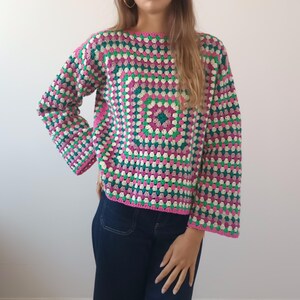 May include: A long-sleeved sweater featuring a multi-colored granny square crochet pattern. The garment is constructed with repeating geometric clusters in shades of vibrant pink, deep forest green, and off-white, finished with a boat neckline.