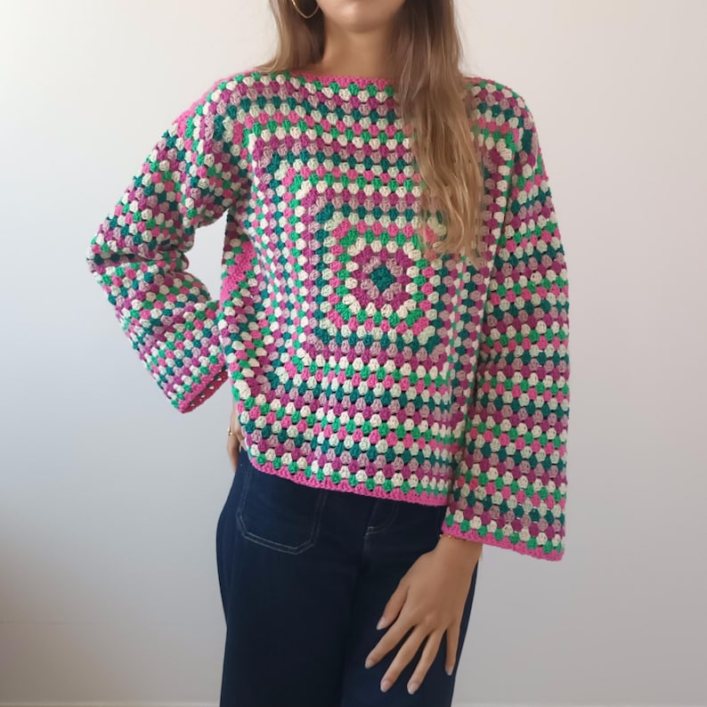 May include: A long-sleeved sweater featuring a multi-colored granny square crochet pattern. The garment is constructed with repeating geometric clusters in shades of vibrant pink, deep forest green, and off-white, finished with a boat neckline.
