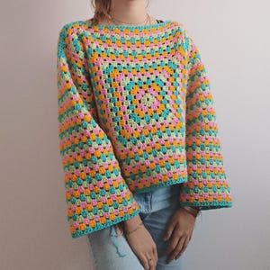 May include: A long-sleeved crochet sweater featuring a vibrant granny square pattern in alternating shades of turquoise, bright pink, and golden yellow. The garment has a wide, boxy fit with loose sleeves and a straight neckline, crafted from yarn.