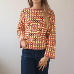 May include: A long-sleeved crochet sweater featuring a multi-colored granny square pattern in shades of pale yellow, burnt orange, and dusty rose. The garment has a wide boat neckline and a straight hem, constructed with a repeating open-stitch design.