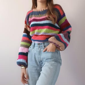 Pode incluir: Um suéter de crochê listrado colorido com mangas compridas e um caimento solto. O suéter apresenta uma variedade de cores, incluindo rosa, azul, verde e marrom.