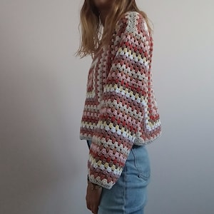 May include: A long-sleeve crochet sweater featuring a multi-colored granny square pattern in shades of dusty rose, cream, light brown, and muted red. The garment has a boxy, cropped silhouette with a wide neckline and ribbed-style cuffs.