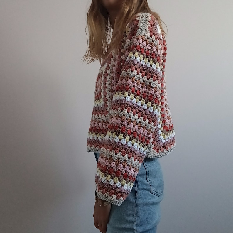 May include: A long-sleeve crochet sweater featuring a multi-colored granny square pattern in shades of dusty rose, cream, light brown, and muted red. The garment has a boxy, cropped silhouette with a wide neckline and ribbed-style cuffs.