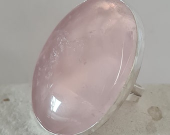 May include: A ring featuring a large, oval-shaped, translucent pale pink rose quartz gemstone set in a smooth, polished silver-toned metal bezel. The stone displays natural internal inclusions and a glossy surface, mounted on a simple silver band.