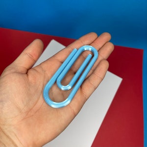 May include: A light blue plastic paperclip. The paperclip is made of a smooth, shiny plastic and is shaped like a traditional paperclip.