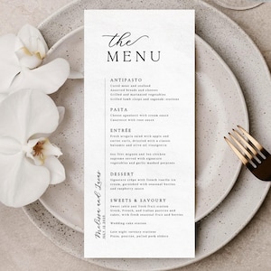 May include: A wedding menu card on a gray plate with a gold fork. The menu features a list of dishes, including antipasto, pasta, entrée, dessert, and sweets. The menu is decorated with an orchid and the names "Melissa and Lucas" and the date "July 12 2025".