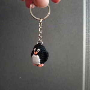 May include: A handmade black and white penguin keychain with orange feet and beak. The small, crocheted penguin is attached to a silver keyring with a chain. The keychain is held up against a gray background.