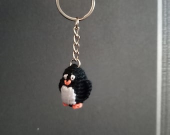 May include: A handmade black and white penguin keychain with orange feet and beak. The small, crocheted penguin is attached to a silver keyring with a chain. The keychain is held up against a gray background.
