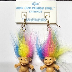 May include: A pair of Good Luck Rainbow Troll earrings. Each earring features a small, tan-colored troll figure with a colorful rainbow-colored hair. The earrings are attached to surgical steel posts and are displayed on a white card with text.
