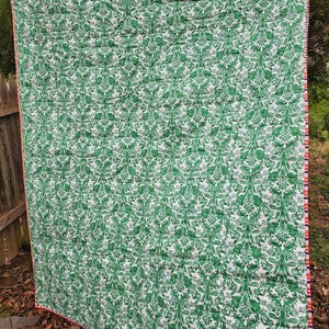 May include: A green and white patterned quilt with a red and white trim. The quilt is folded and lying on the ground.