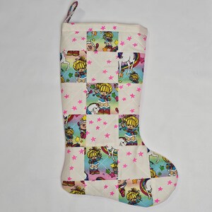 May include: A quilted Christmas stocking with a patchwork design. The fabric features a repeating pattern of a cartoon girl, unicorns, and rainbows. Pink stars are scattered across the white background. The stocking has a pink loop for hanging.