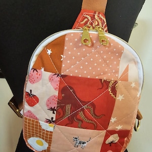May include: A small, quilted crossbody bag with a patchwork design. The bag features a variety of patterns, including strawberries, an octopus, eggs, and stars. The bag has a zipper closure and a brown strap.