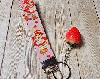 May include: A pink and white fabric keychain with a strawberry print and a strawberry charm. The keychain features a repeating pattern of strawberries and a cartoon character.