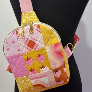 K&ouml;nnte beinhalten: Eine Patchwork-Crossbody-Tasche in einem gelben, rosa und roten Farbschema. Die Tasche verf&uuml;gt &uuml;ber eine Vielzahl von Mustern, darunter Erdbeeren, Karos und Blumenmuster. Die Tasche hat einen Rei&szlig;verschluss und einen goldfarbenen Rei&szlig;verschluss. Die Tasche ist aus gestepptem Stoff gefertigt.